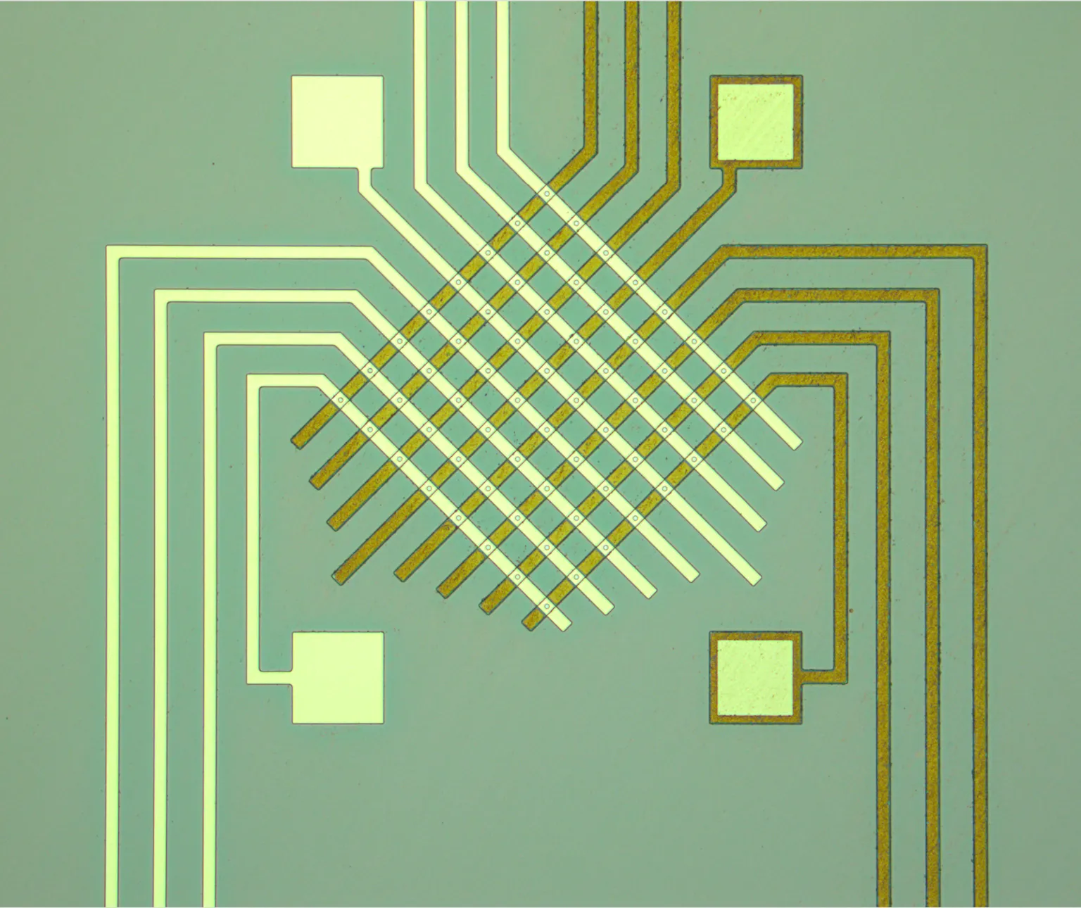 A brightfield microscope image of a fabricated memristor crossbar: cream and gold electrode traces crossing diagonally on a pale green substrate, with four square bond pads at the corners.