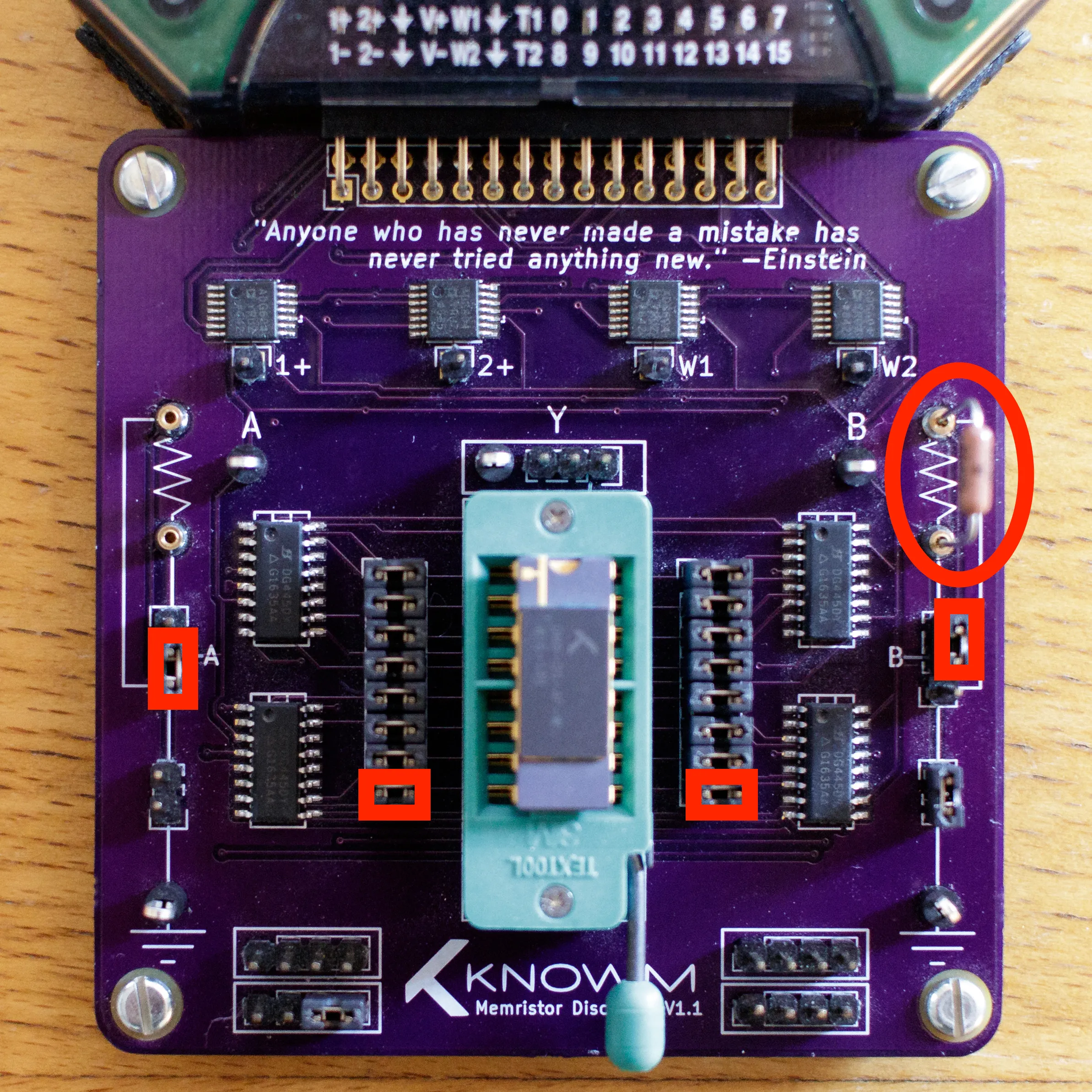 Photo of the purple Knowm Memristor Discovery V1.1 board on a wooden desk: a chip socket in the center, banks of analog switches and muxes around it, an Einstein quote silkscreened across the top, and red annotations marking the jumper and series-resistor configuration.
